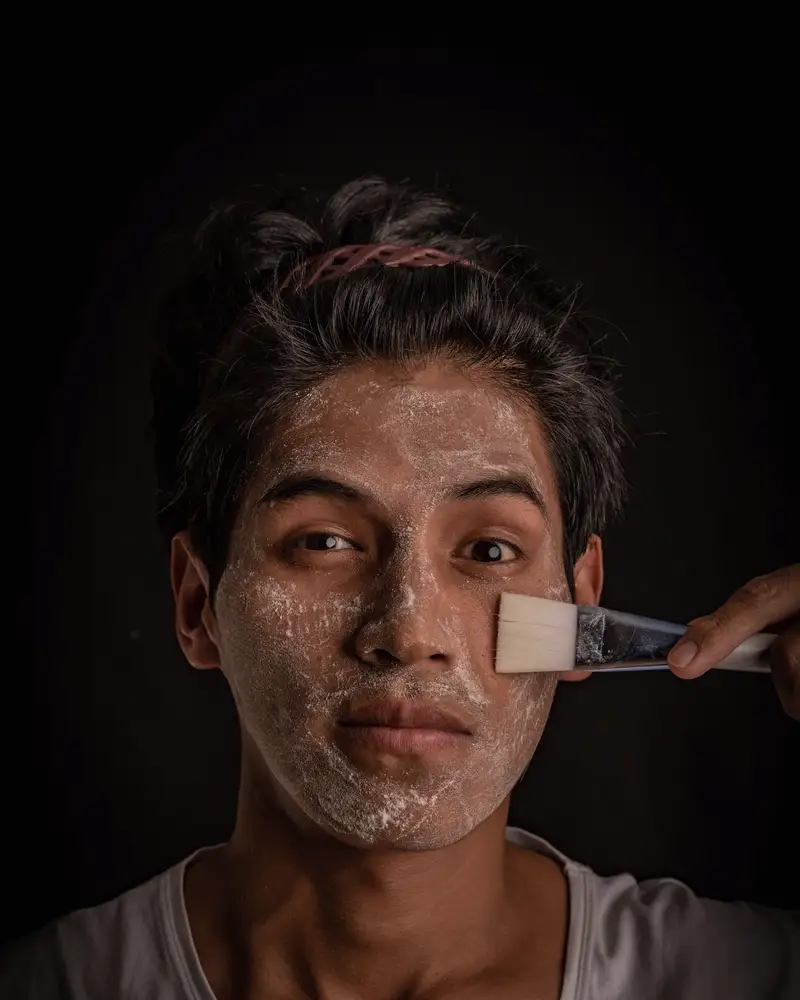 Man with a clay treatment mask being applied by brush, lit from one side against a dark wall